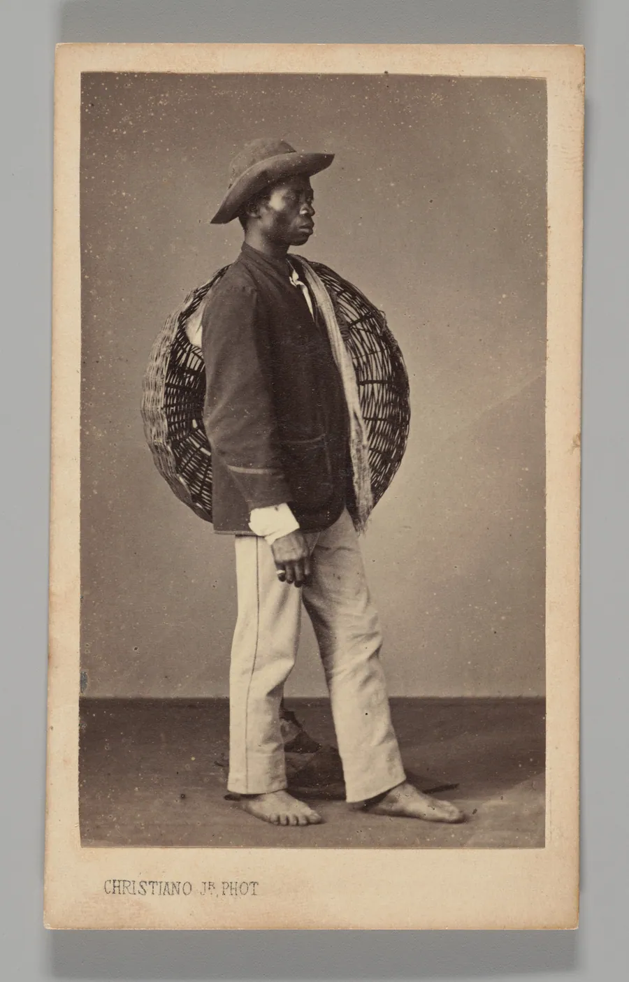 [Studio Portrait: Male Street Vendor Standing in Profile Holding Basket, Brazil] by Georges Leuzinger